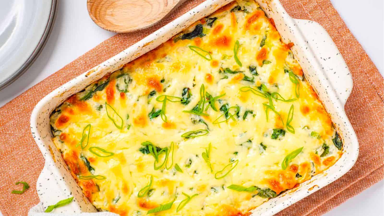A baked casserole dish filled with a cheesy spinach and egg mixture, garnished with sliced green onions, placed on an orange cloth beside a wooden spoon.