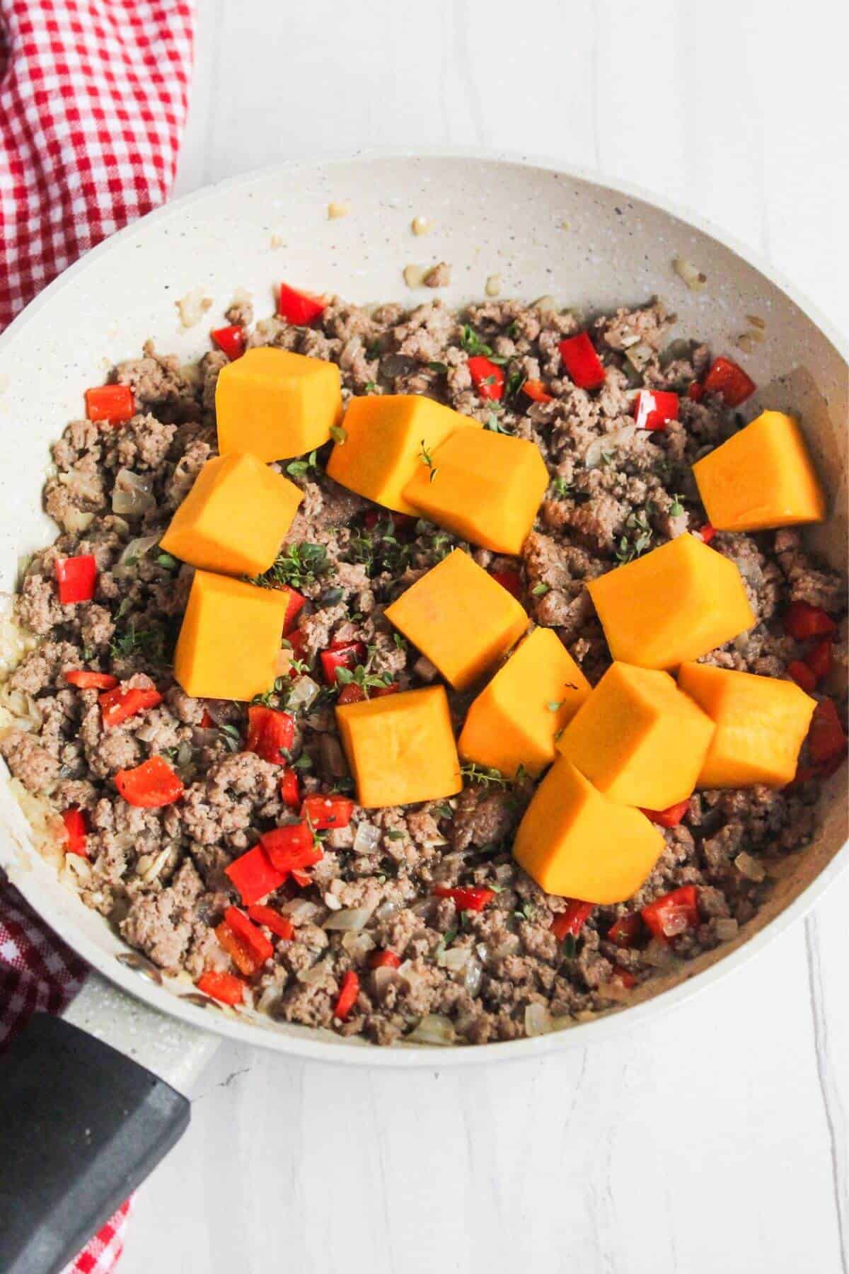 A skillet with ground turkey, red bell peppers, onions, and chunks of butternut squash rests on a white surface. A red and white checkered cloth is partially visible.