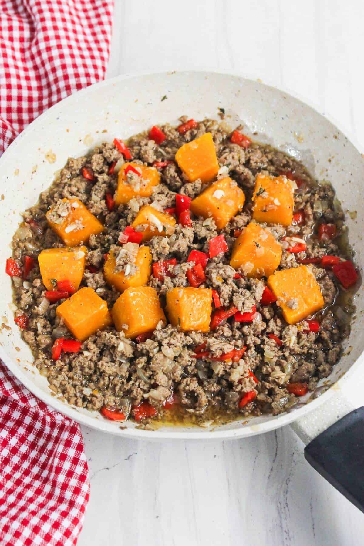 A white skillet with ground turkey, cubed orange butternut squash, and chopped red bell peppers sits elegantly on a white surface beside a red and white checkered cloth.