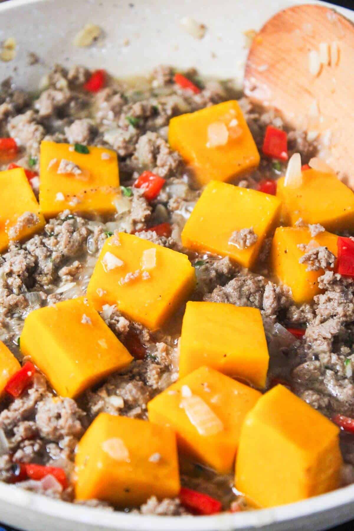 A pan with ground turkey, diced red peppers, chunks of butternut squash, and onions being cooked. A wooden spoon is visible at the side.