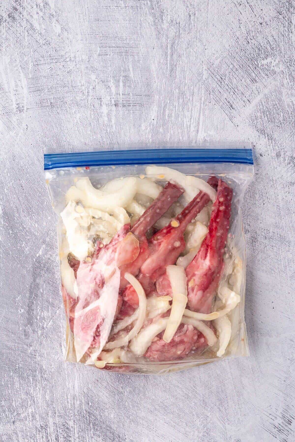 Raw marinated meat and sliced onions in a clear ziplock bag on a textured gray surface.