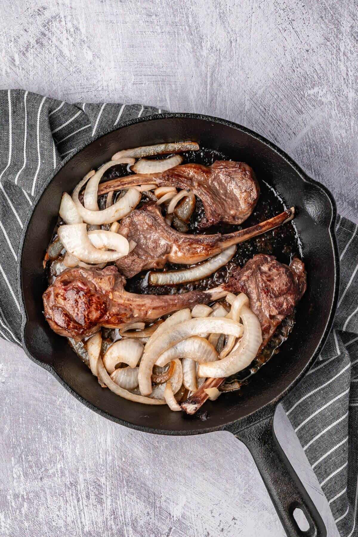 Cast iron skillet with cooked lamb chops and sliced onions on a striped cloth.