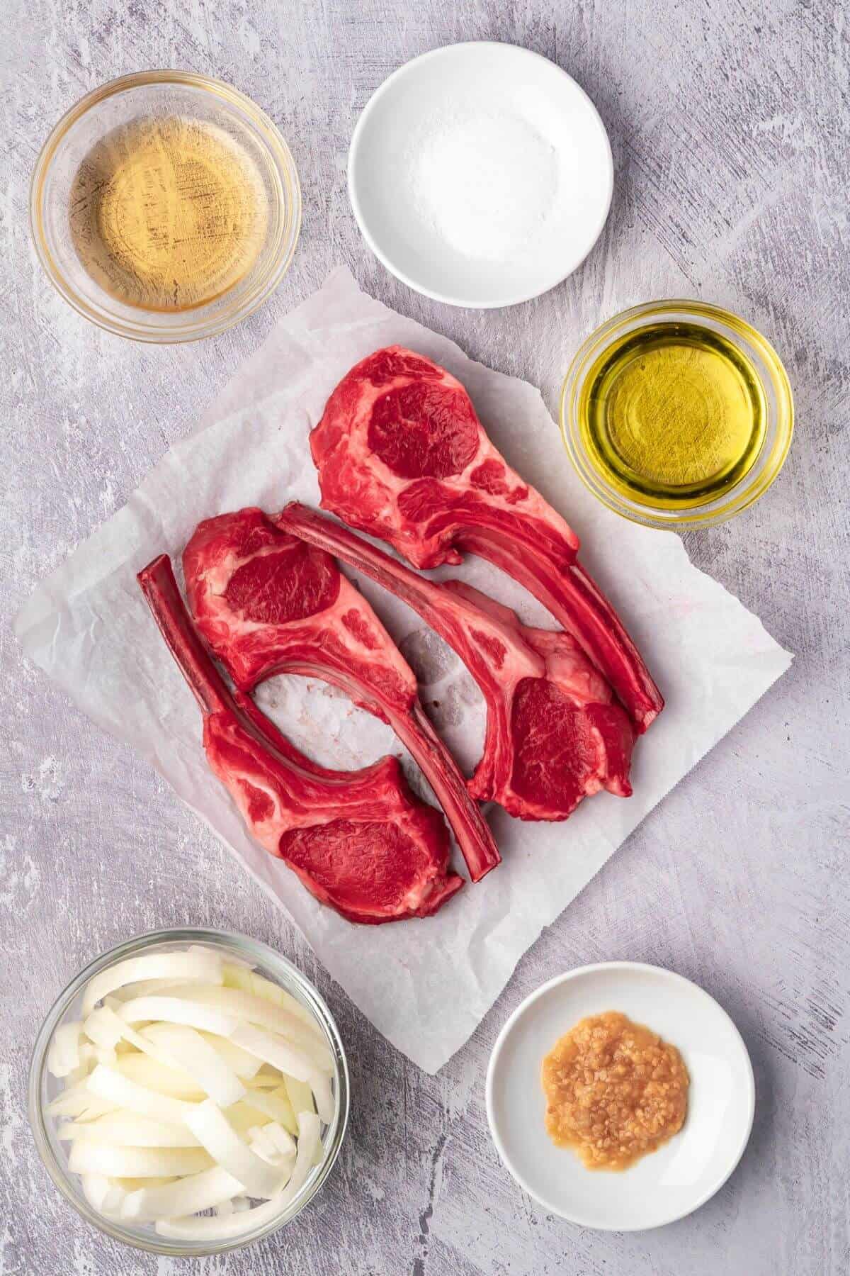Raw lamb chops on parchment paper surrounded by bowls of oil, vinegar, salt, sliced onions, and minced garlic on a gray surface.