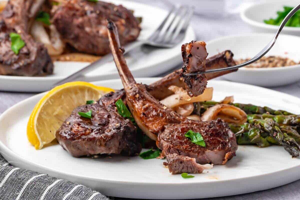 Grilled lamb chops with herbs and lemon wedge on a white plate, accompanied by asparagus and onions. Fork lifting a piece of lamb.