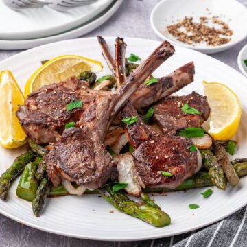 Lamb chops garnished with parsley on a plate with asparagus and lemon wedges.