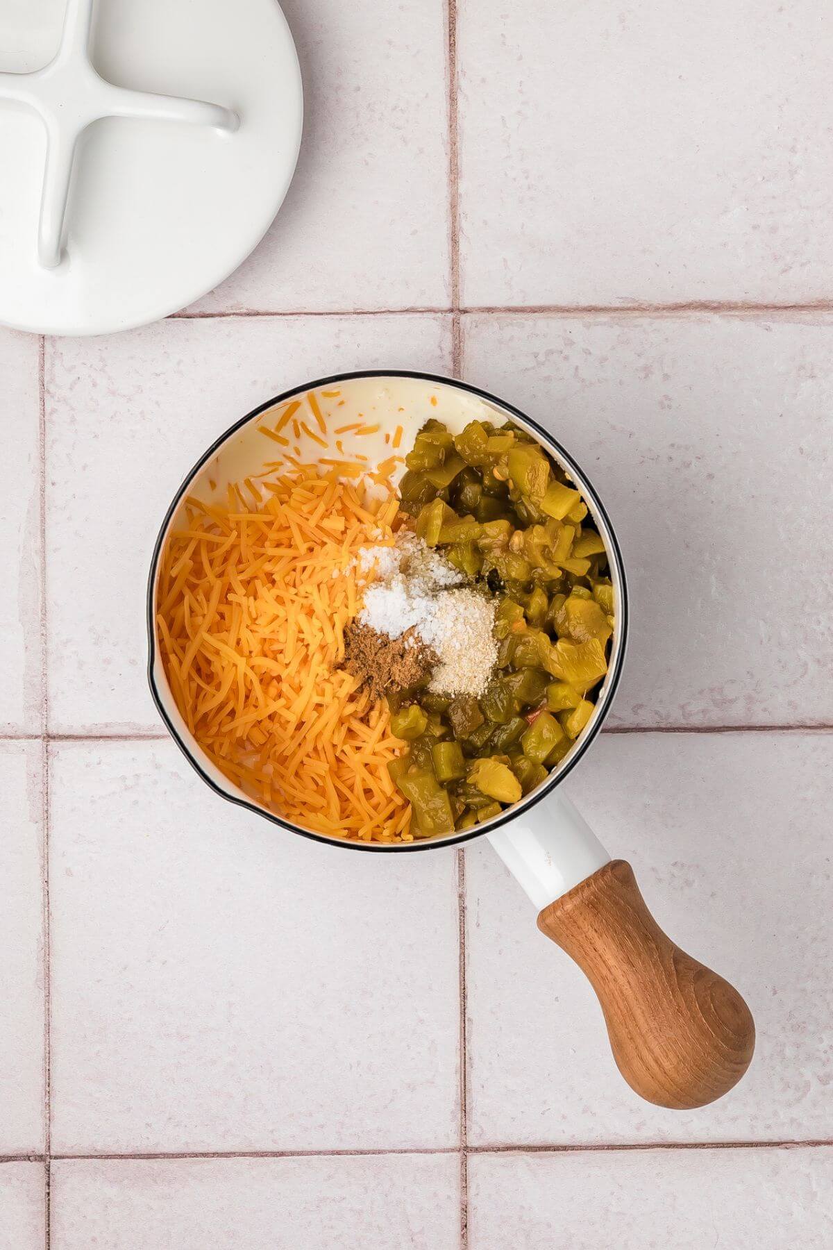 A saucepan on a tiled surface contains shredded cheese, chopped green chiles, and spices, ready for mixing.