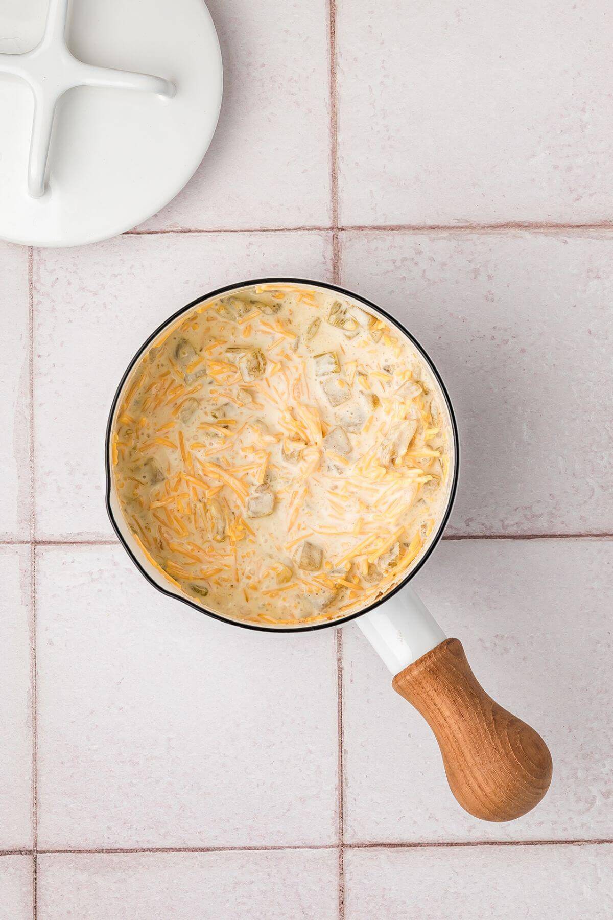 Small pot with a wooden handle containing melted cheese and diced green chiles on a tiled surface. Lid partially visible in the corner.