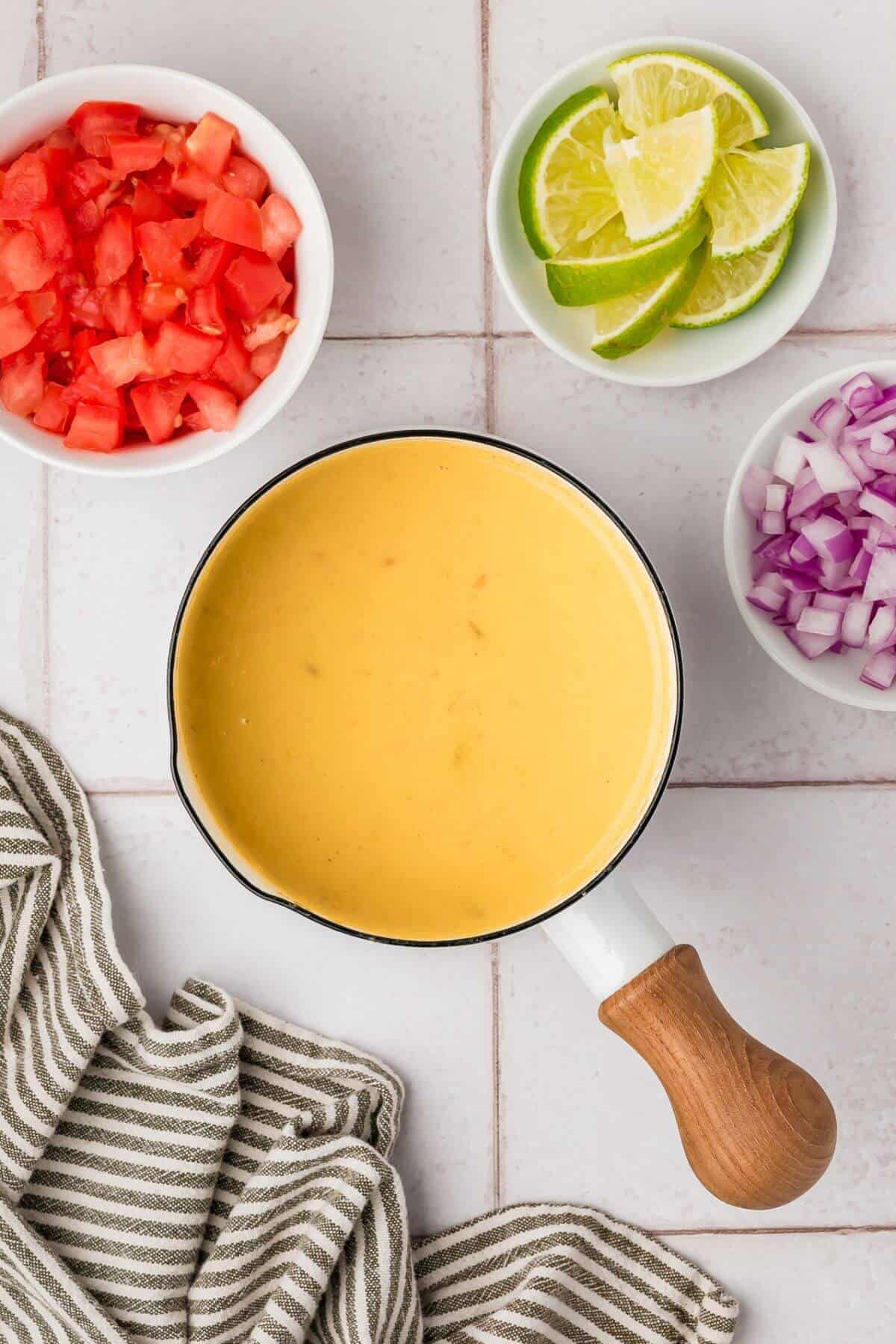 A pot of yellow queso dip is centered on a table, surrounded by bowls of chopped tomatoes, lime wedges, and diced red onions. A striped cloth is placed nearby.