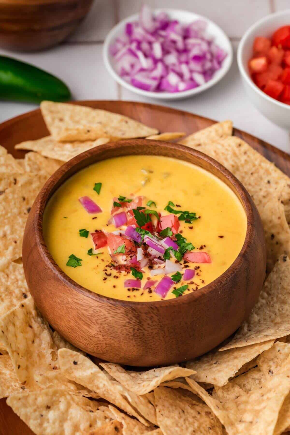 A wooden bowl of cheese dip garnished with chopped onions, tomatoes, and herbs, surrounded by tortilla chips.
