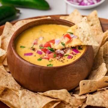 A wooden bowl filled with cheesy dip, topped with diced tomatoes, onions, and cilantro, surrounded by tortilla chips.