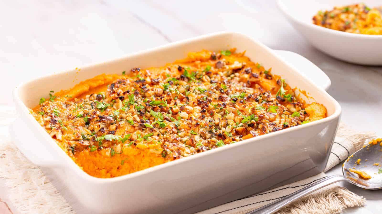 Baked sweet potato casserole with a crispy nut topping in a rectangular dish, garnished with herbs.