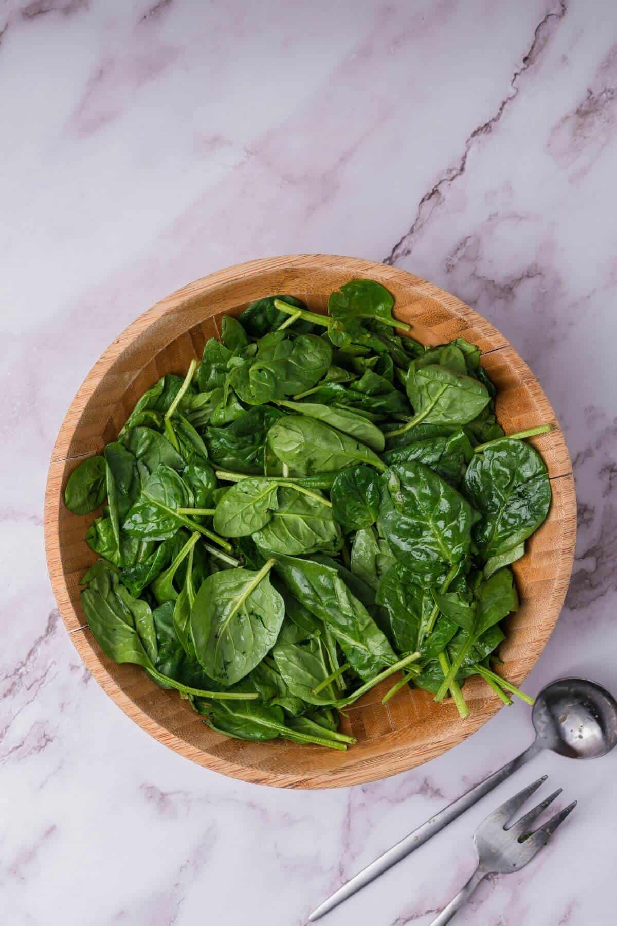 A wooden bowl filled with fresh spinach leaves on a marble countertop, next to a metal serving spoon and fork.
