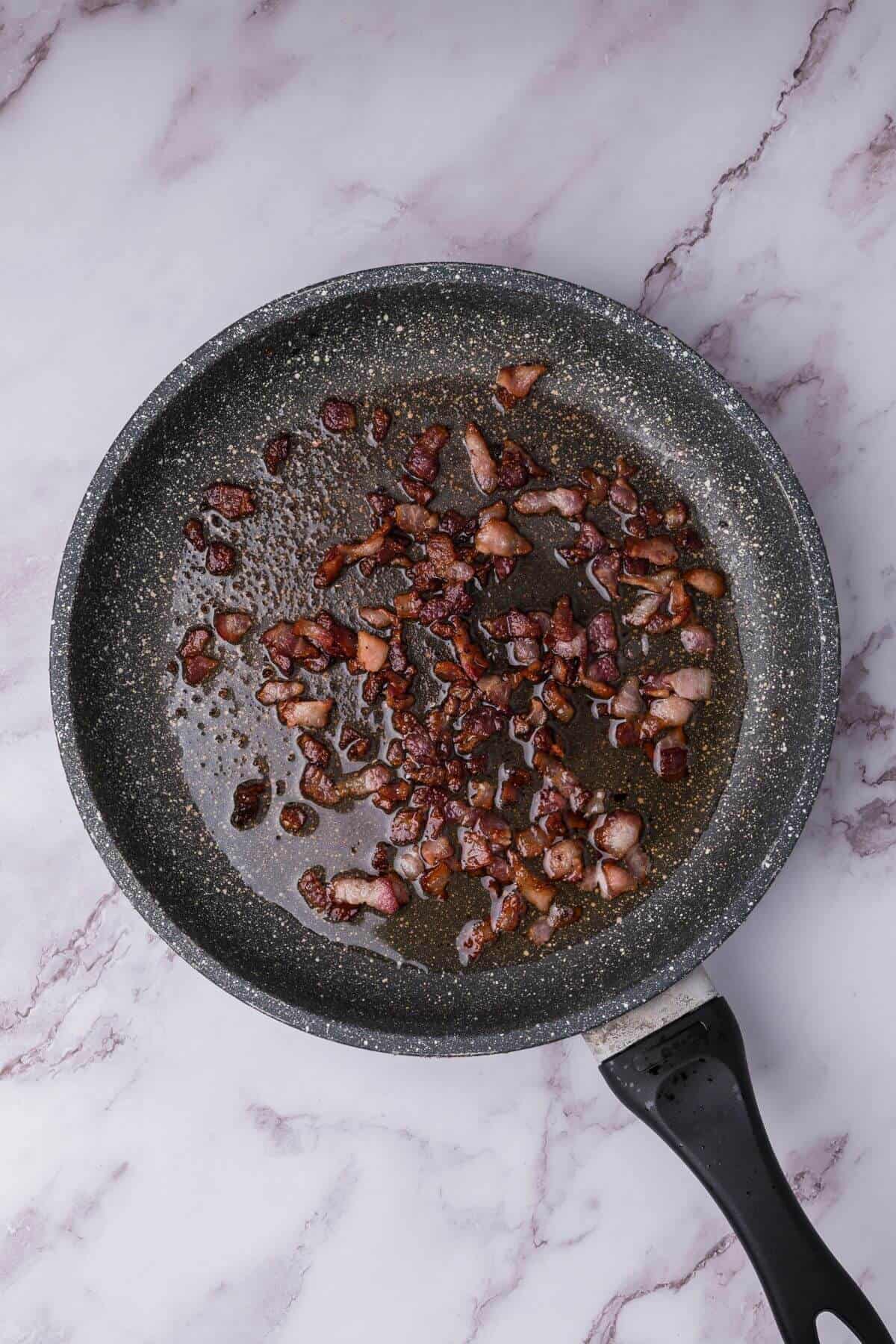 Frying pan with cooked bacon pieces on a marble countertop.