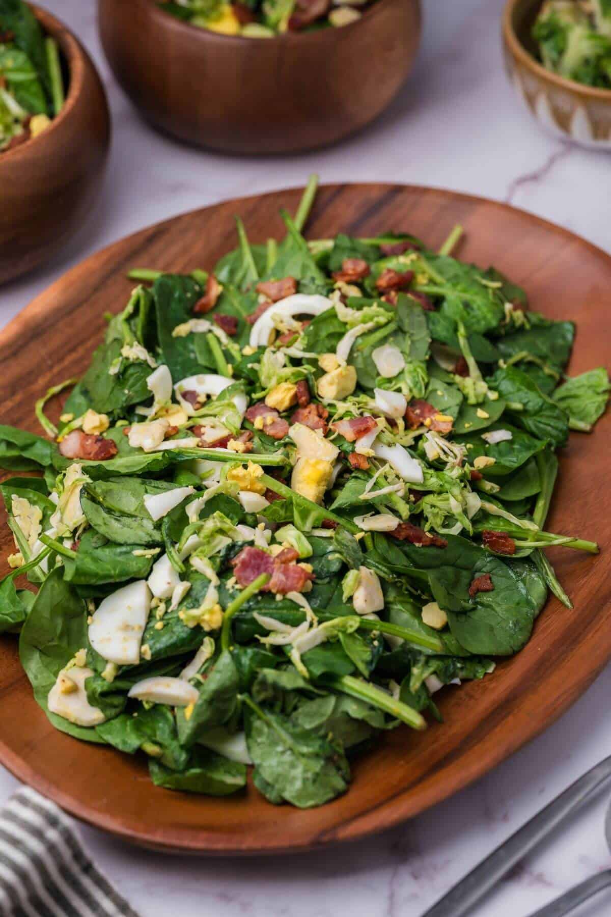A wooden plate with a fresh green salad topped with chopped boiled eggs, bacon bits, and possibly cheese pieces.