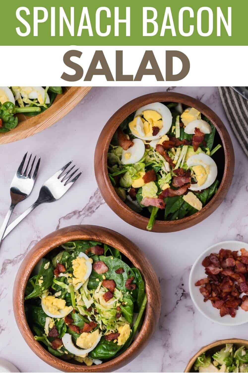 Bowls of spinach salad with bacon, boiled eggs, and shredded cheese on a marble table, accompanied by two forks and a small dish of bacon bits.