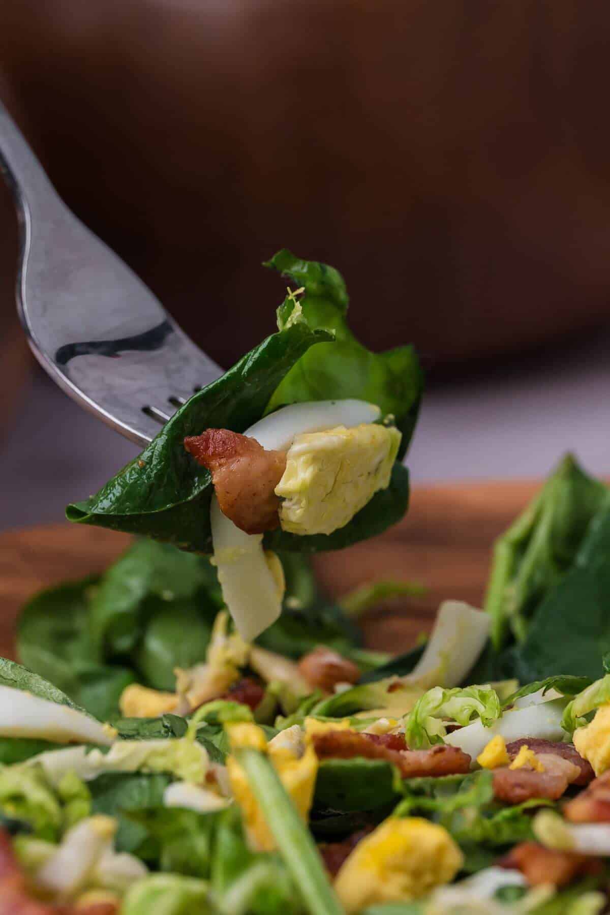 Close-up of a fork holding a salad with spinach, bacon, hard-boiled egg, and avocado over a wooden bowl containing more of the salad mix.
