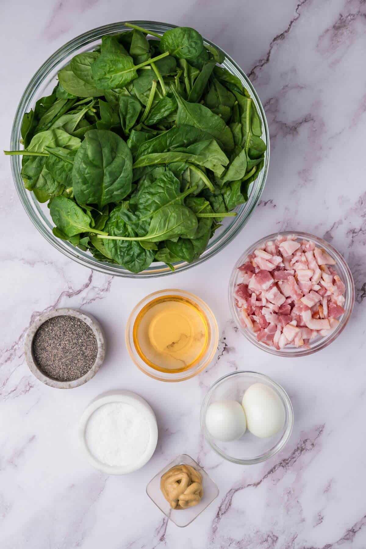 A bowl of spinach, a bowl of chopped bacon, two boiled eggs, small bowls of mustard, salt, and black pepper, and a small dish of oil on a marble countertop.