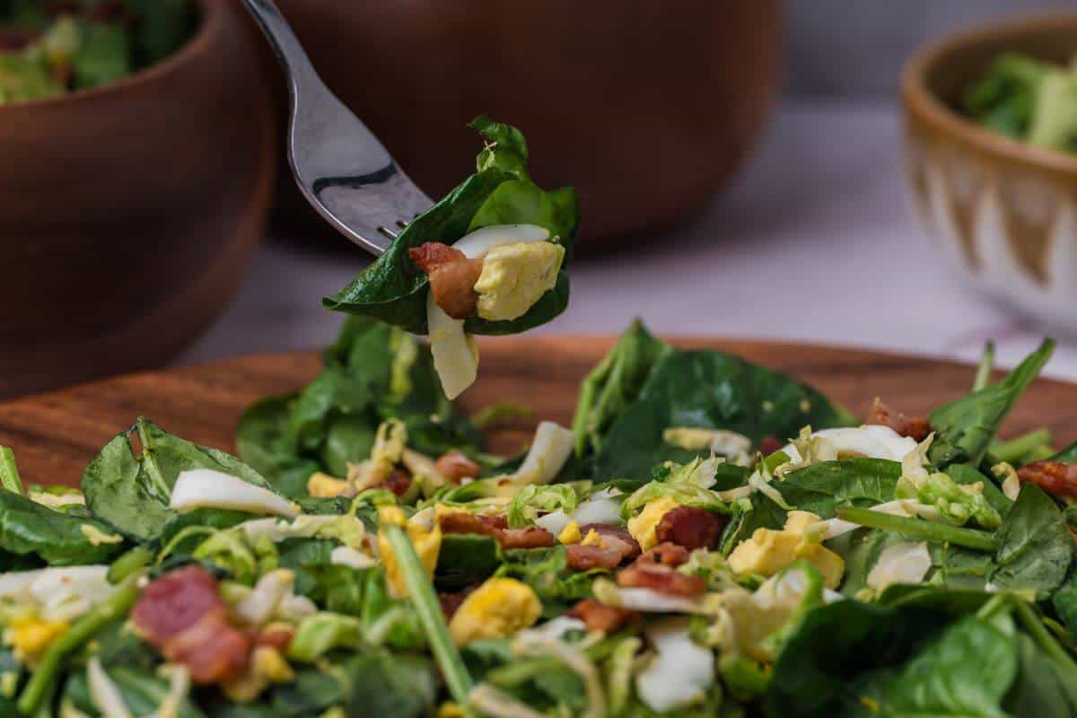 Close-up of a salad featuring spinach, chopped boiled egg, bacon bits, and shredded cabbage on a wooden plate, with a fork lifting a bite.