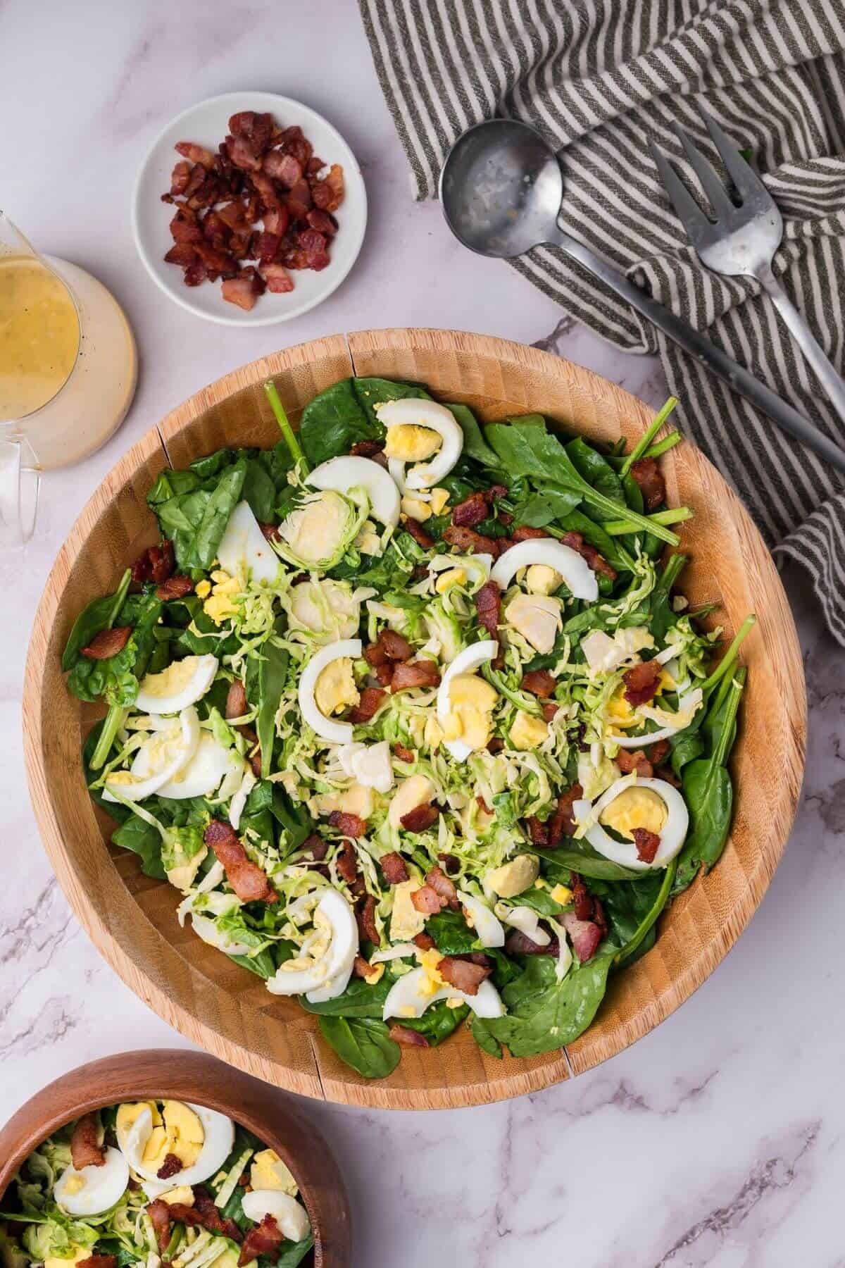 A wooden bowl of salad with spinach, sliced hard-boiled eggs, bacon bits, and shredded vegetables, accompanied by dressing, a small bacon dish, striped cloth, and metal utensils.