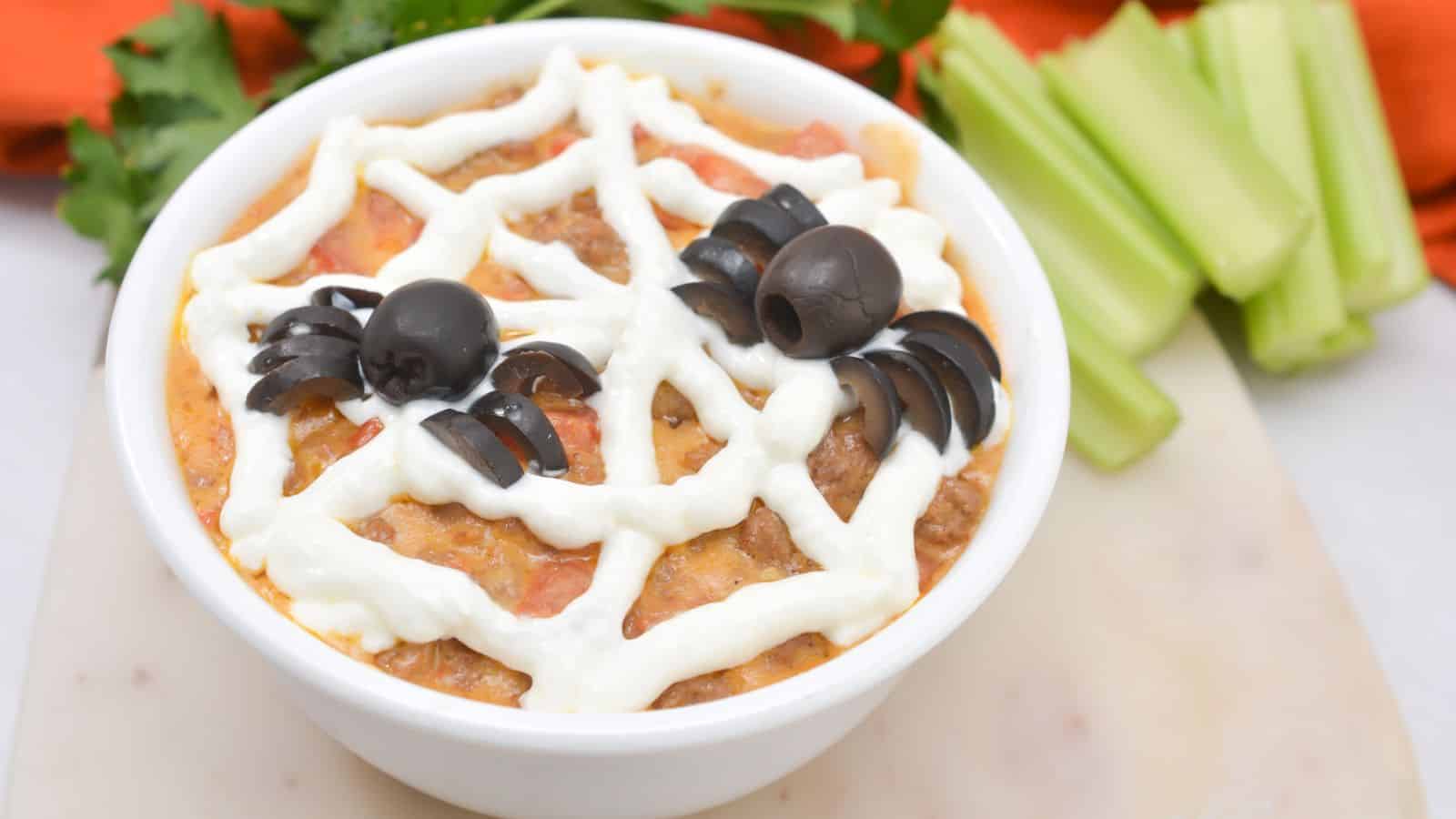 A bowl of dip topped with a spider web design made of sour cream and olive spiders, placed on a marble board with parsley, celery, and an orange napkin nearby.