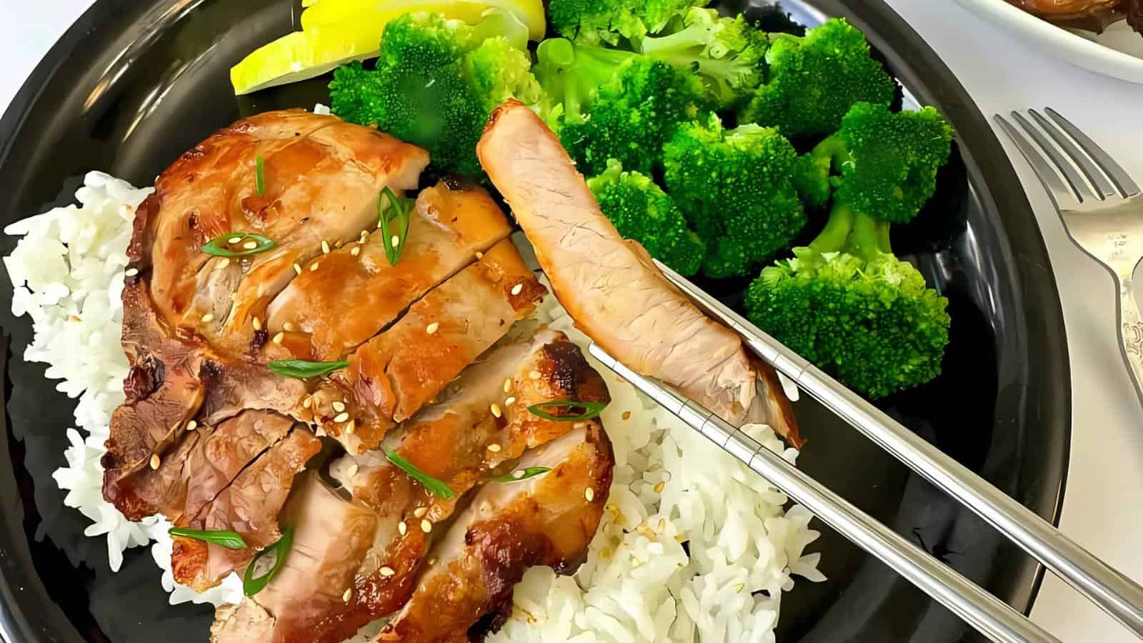 Grilled chicken slices on white rice, garnished with sesame seeds and spring onion, served with steamed broccoli and lemon slices on a black plate.
