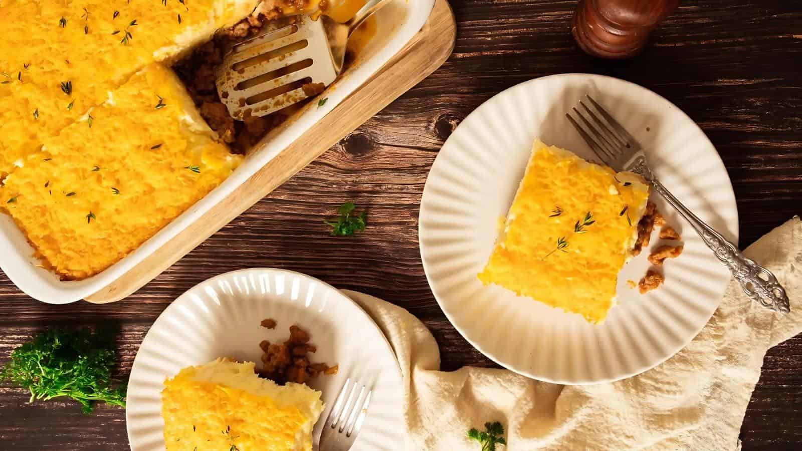 Two plates with servings of shepherd's pie beside a dish. A spatula rests in the main dish. Parsley is scattered on a wooden table, with a beige cloth nearby.