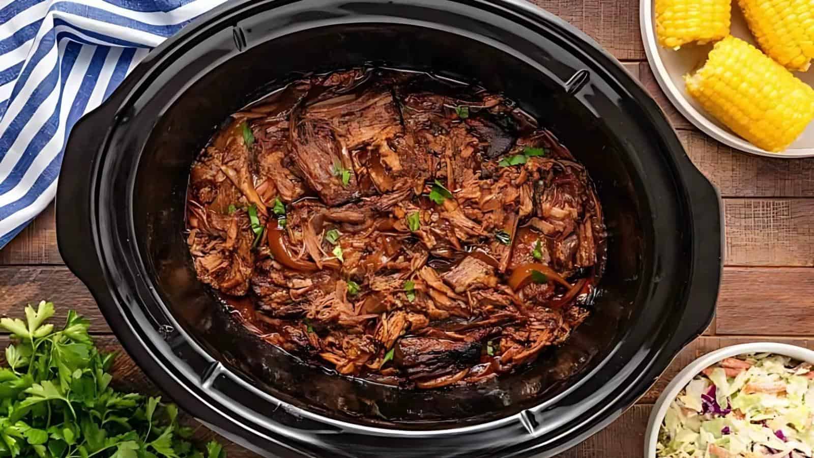 Shredded beef in a black slow cooker, garnished with herbs. Nearby are dishes of corn on the cob and coleslaw.