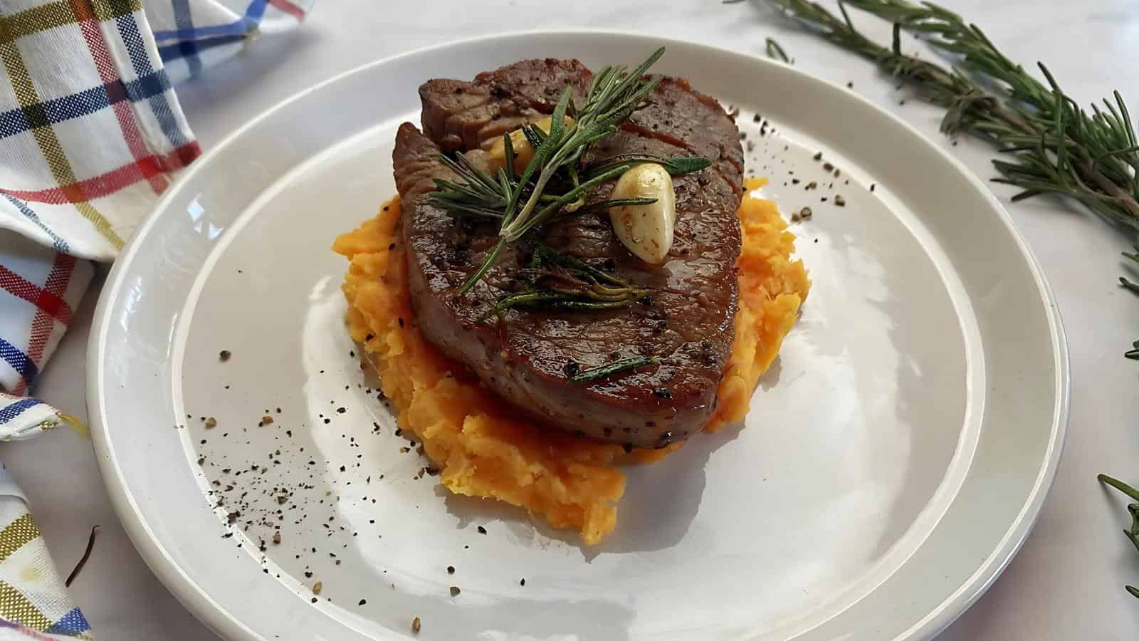 Seared steak topped with garlic and rosemary on a bed of mashed sweet potatoes, garnished with pepper, on a white plate.