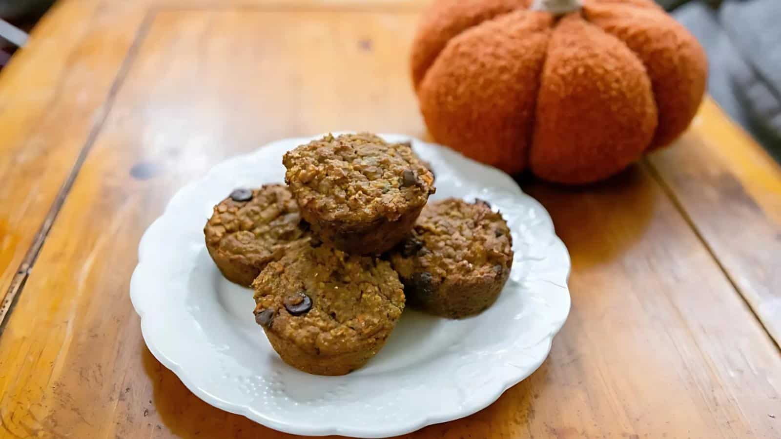 Four chocolate chip muffins on a white plate next to a knitted pumpkin decoration on a wooden table.