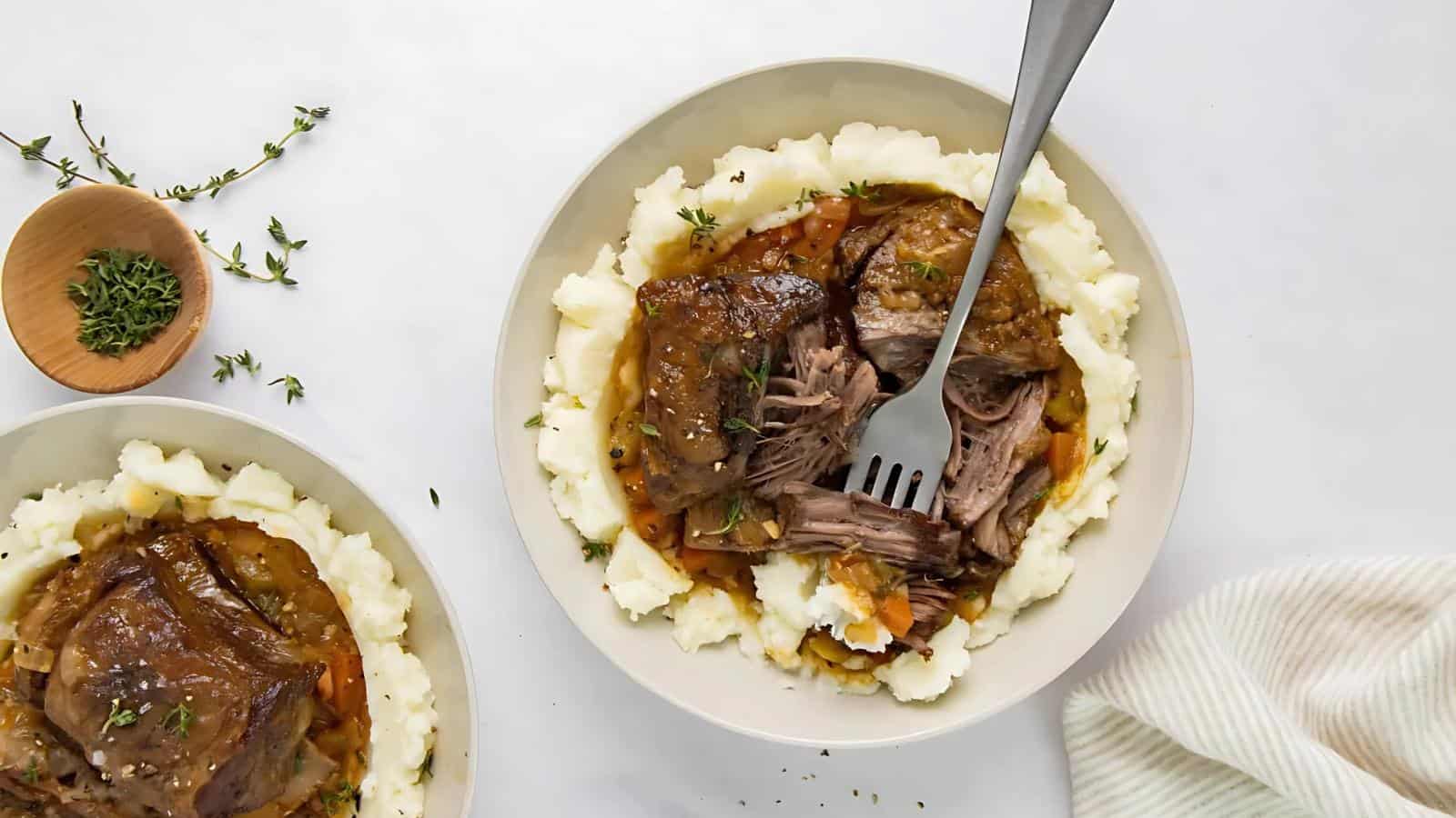 Plate of mashed potatoes topped with shredded beef and gravy. A fork is on the dish. A small bowl of herbs is nearby.