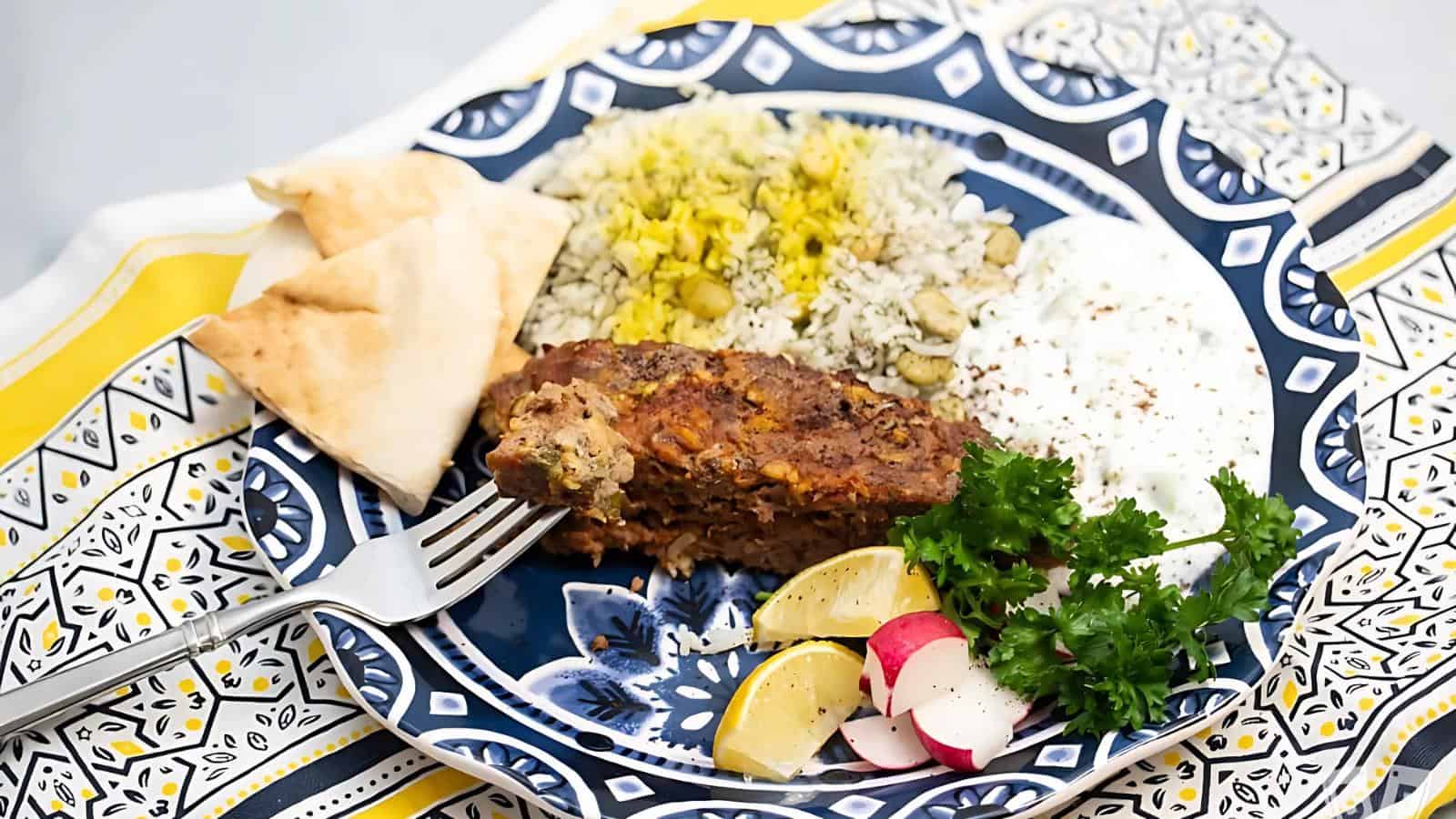 A plate with rice, a portion of meatloaf, radish slices, lemon wedges, pita bread, and a dollop of yogurt garnished with parsley on a patterned plate.