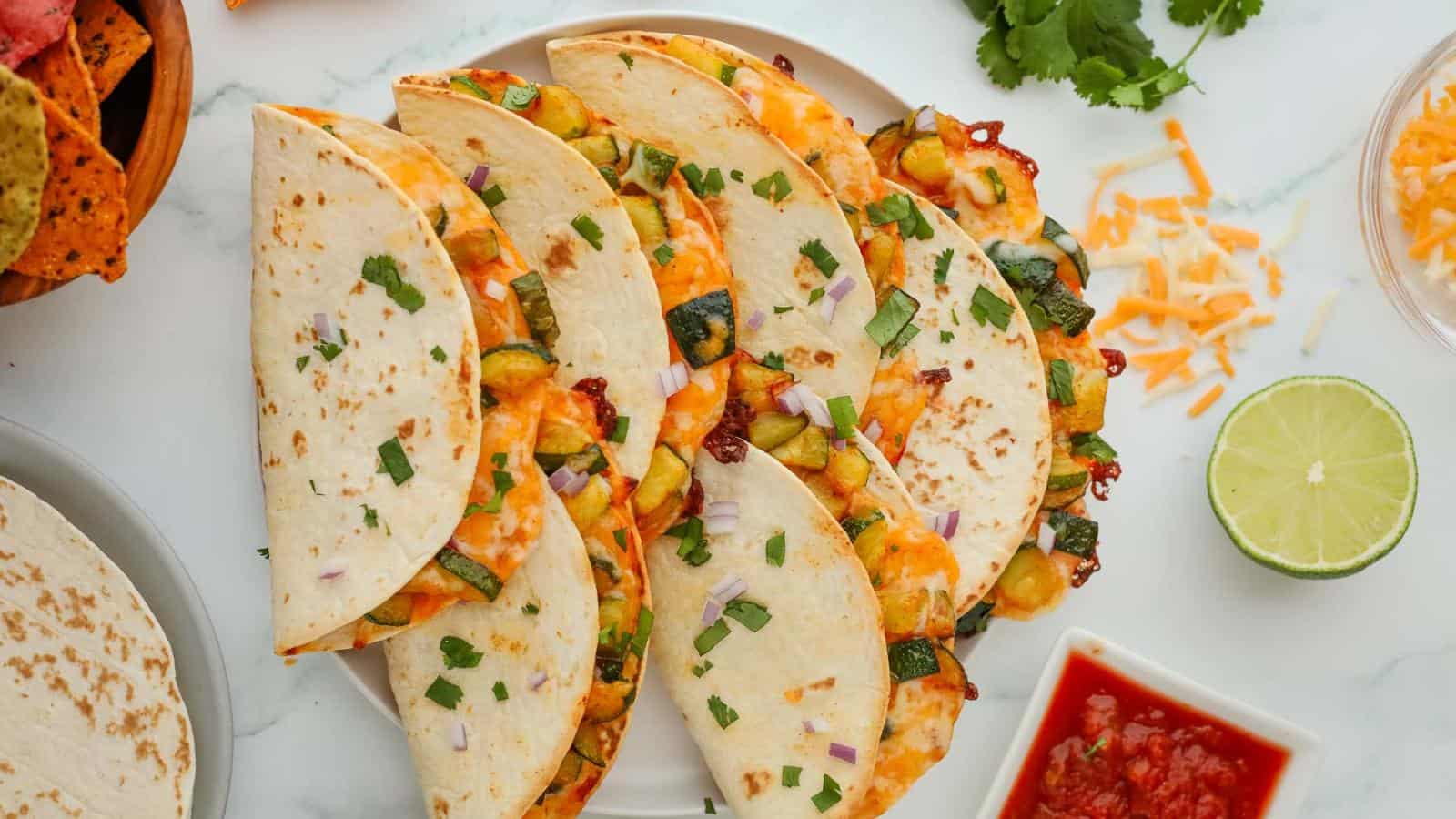 Plate of cheese and vegetable quesadillas garnished with chopped herbs, surrounded by lime, salsa, and shredded cheese on a marble surface.