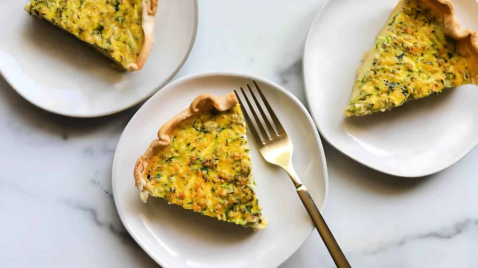 Three slices of vegetable quiche are served on white plates, each with a fork beside them. The crust is golden, and the filling appears seasoned with herbs.