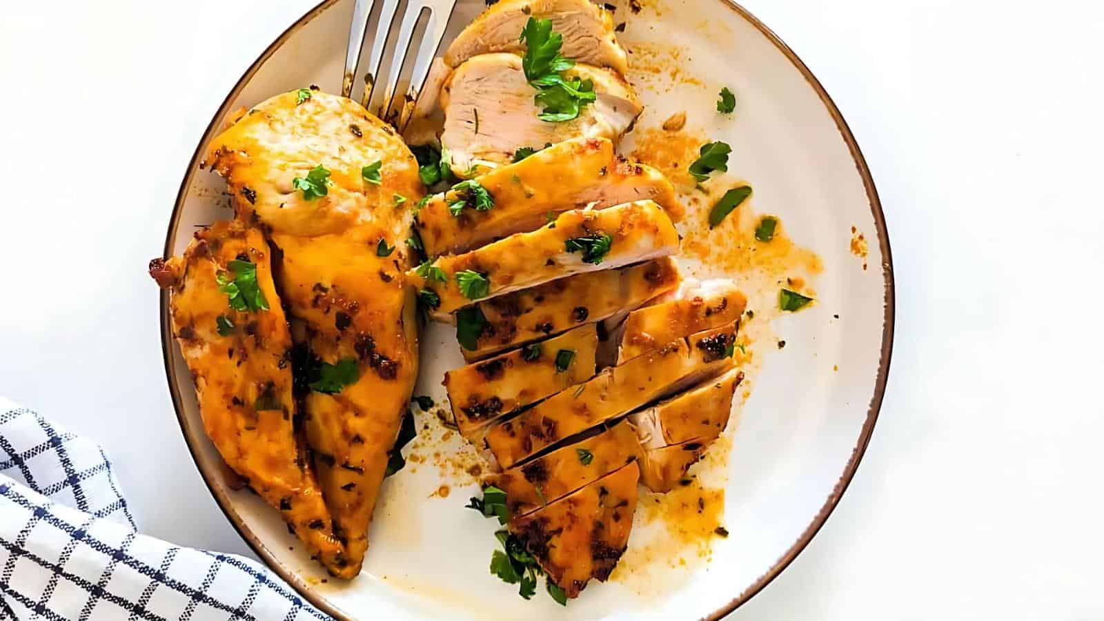 A plate of grilled chicken garnished with green herbs. A fork rests beside the chicken, and a checkered cloth is partially visible in the foreground.