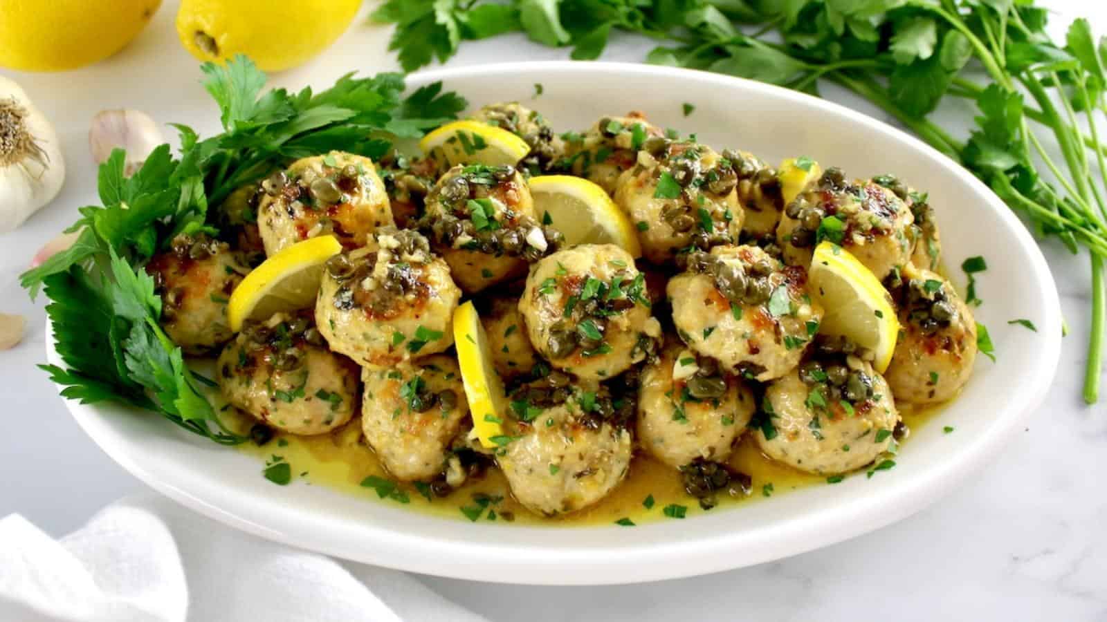 A plate of chicken meatballs garnished with lemon wedges and herbs, topped with a caper sauce, and surrounded by parsley leaves.