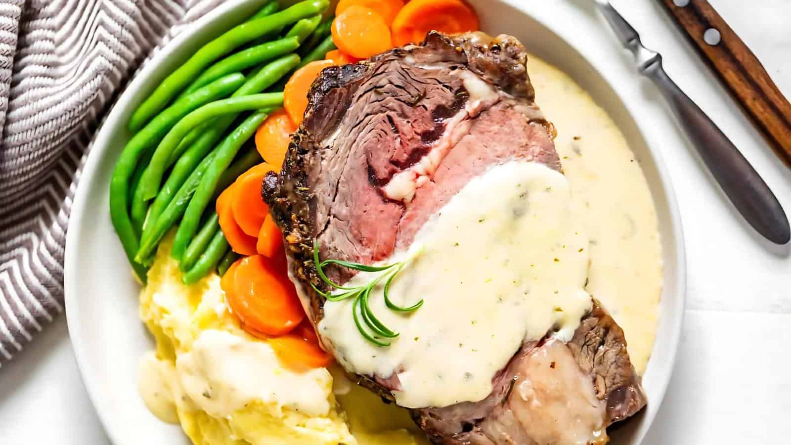 A plate with roast beef topped with white sauce, mashed potatoes, green beans, and sliced carrots, garnished with rosemary. A fork and knife are on the side.