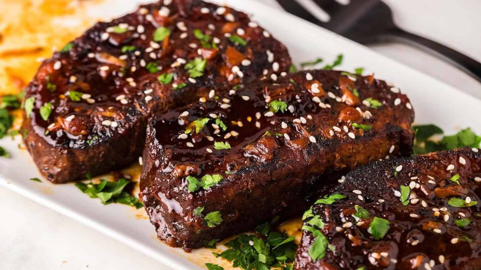 Three glazed beef steaks garnished with sesame seeds and chopped herbs on a white rectangular plate.