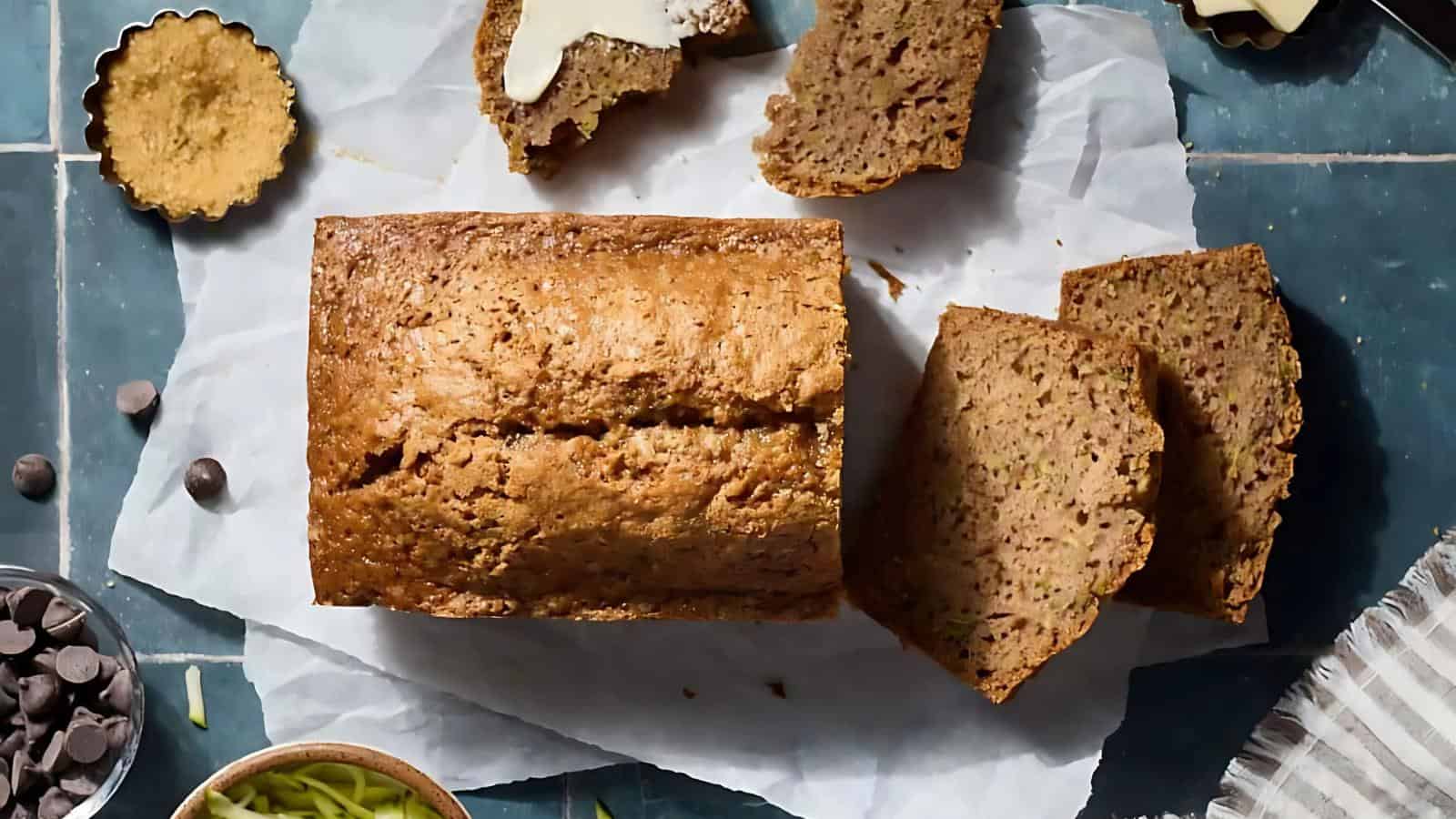 A loaf of bread on parchment paper, partially sliced, with chocolate chips and green shreds in bowls and scattered. A slice is buttered, and baking utensils are nearby.