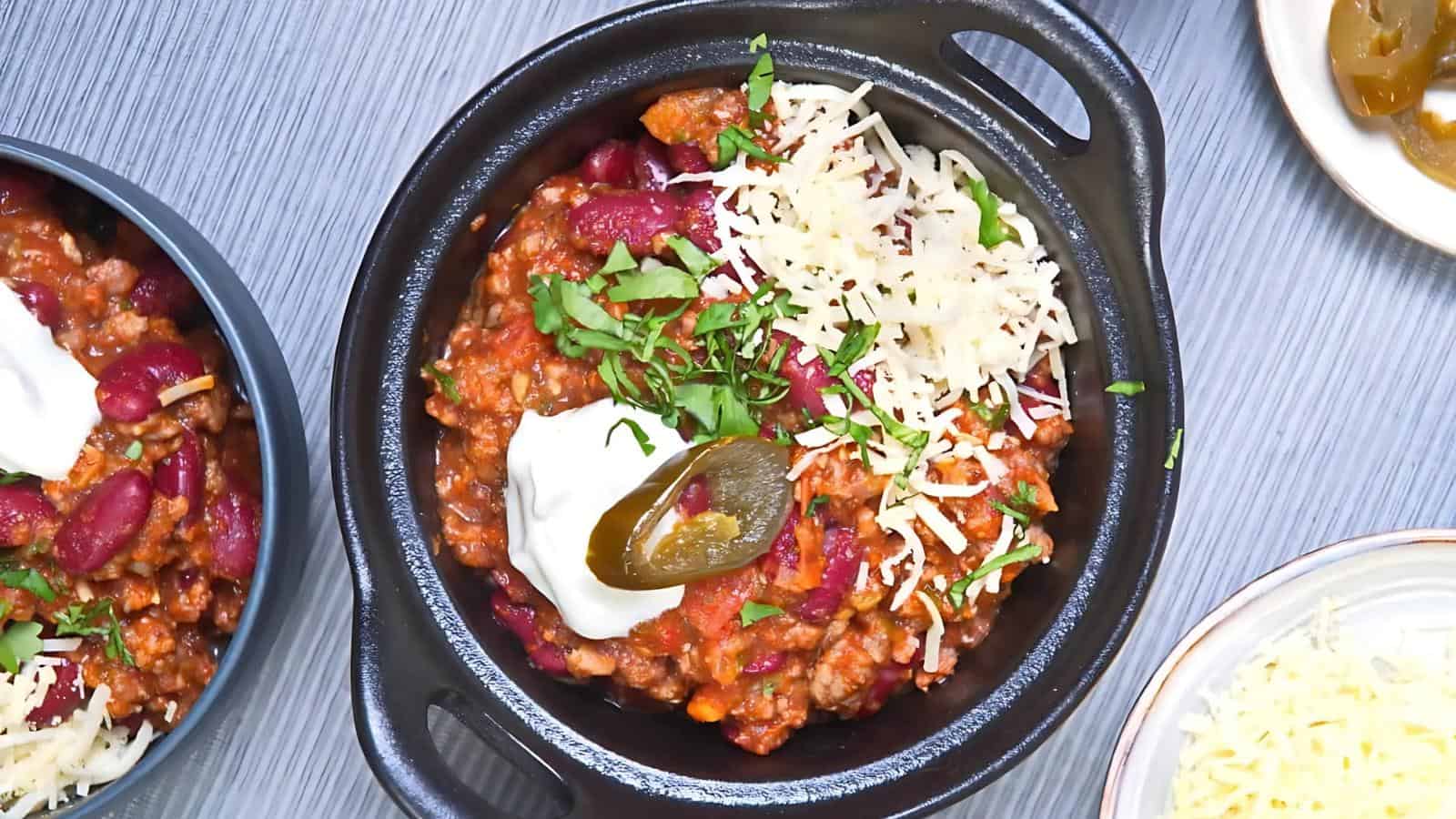 Bowl of chili con carne with kidney beans, topped with sour cream, shredded cheese, jalapeño, and chopped herbs.