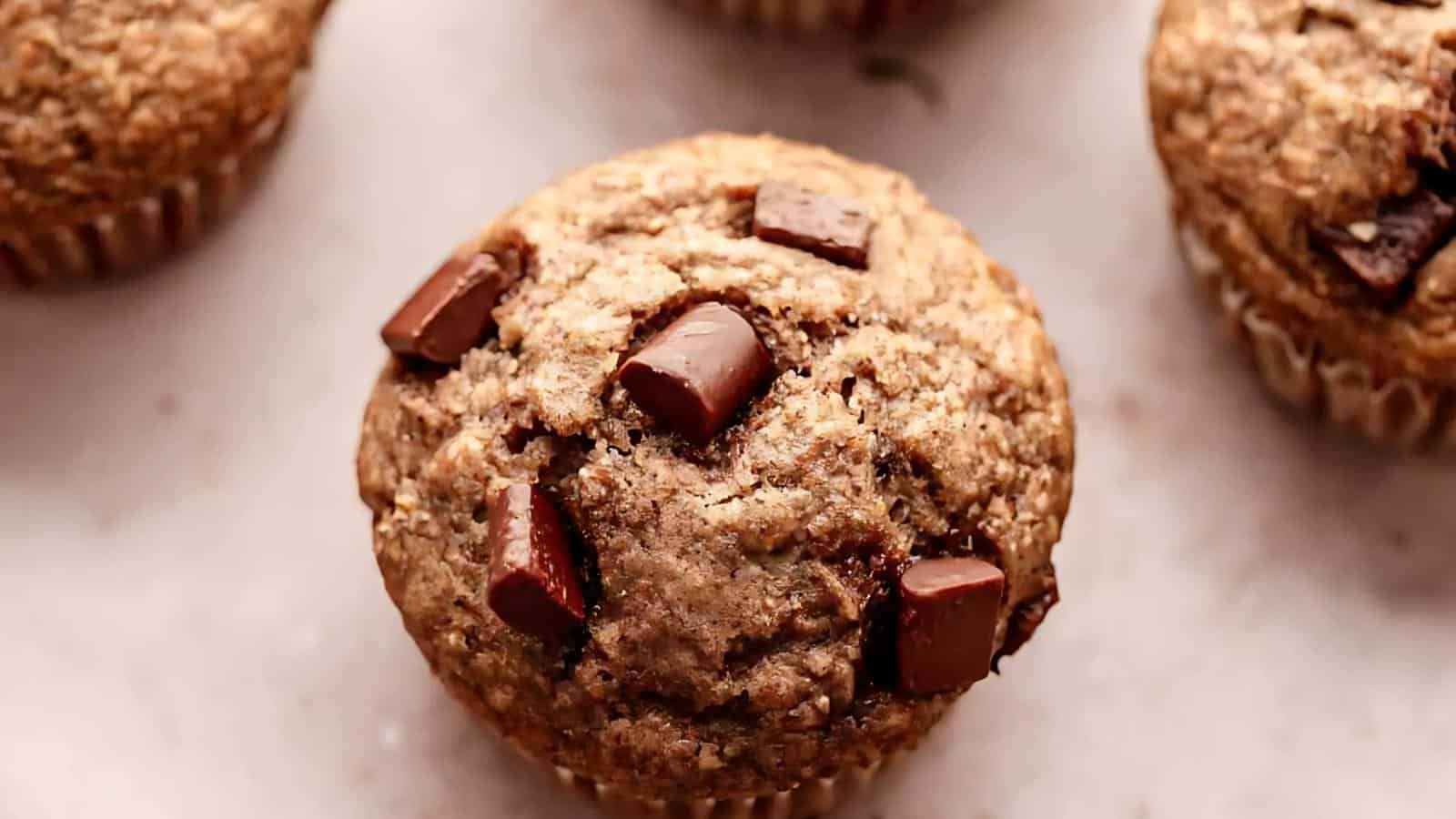 A close-up of a chocolate chip muffin, featuring a golden-brown crumb and visible chunks of chocolate, on a light surface.