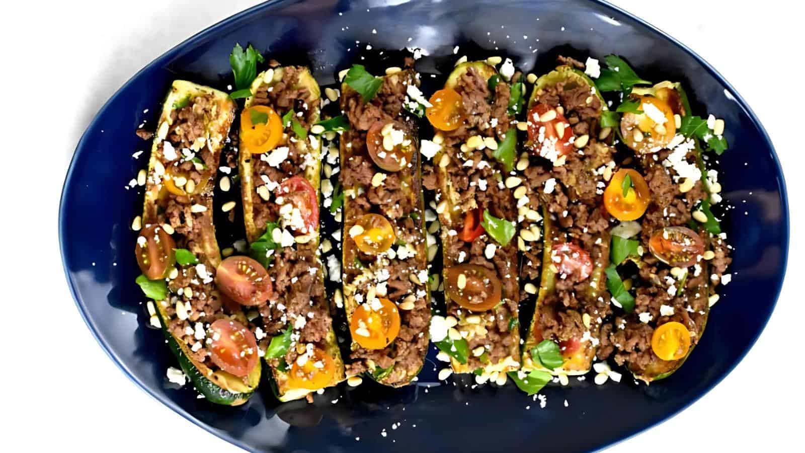 Stuffed zucchini boats topped with ground meat, cherry tomatoes, herbs, and crumbled cheese on a dark blue oval plate.