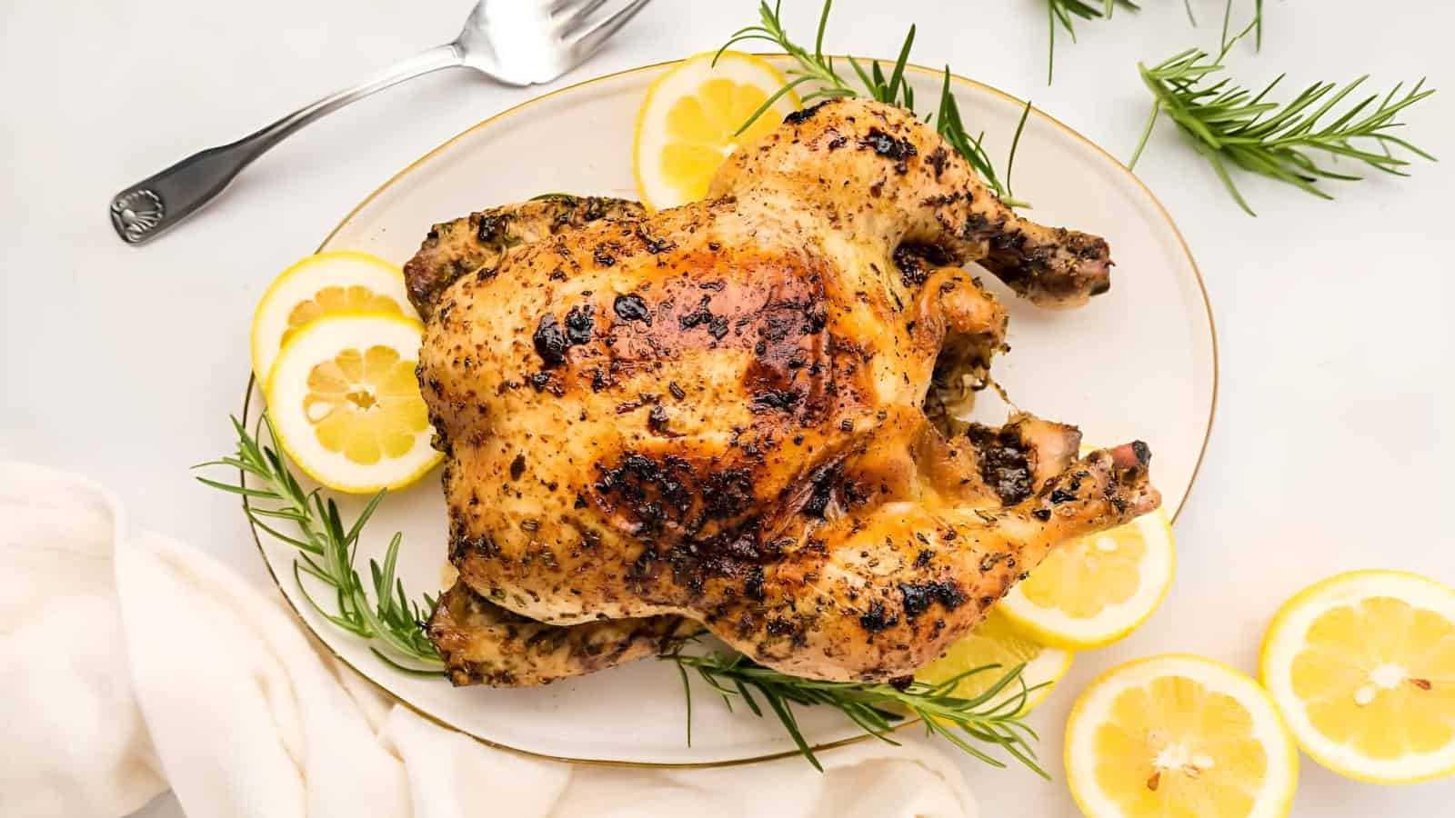 Roasted chicken garnished with lemon slices and rosemary on a white plate, with a fork and napkin nearby.