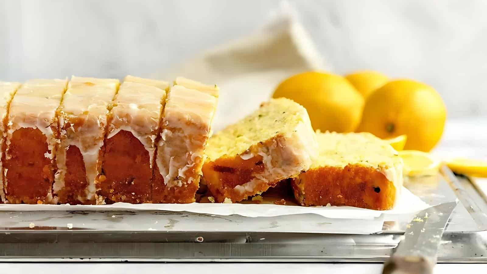 Sliced lemon cake with icing on parchment paper, accompanied by whole lemons in the background.