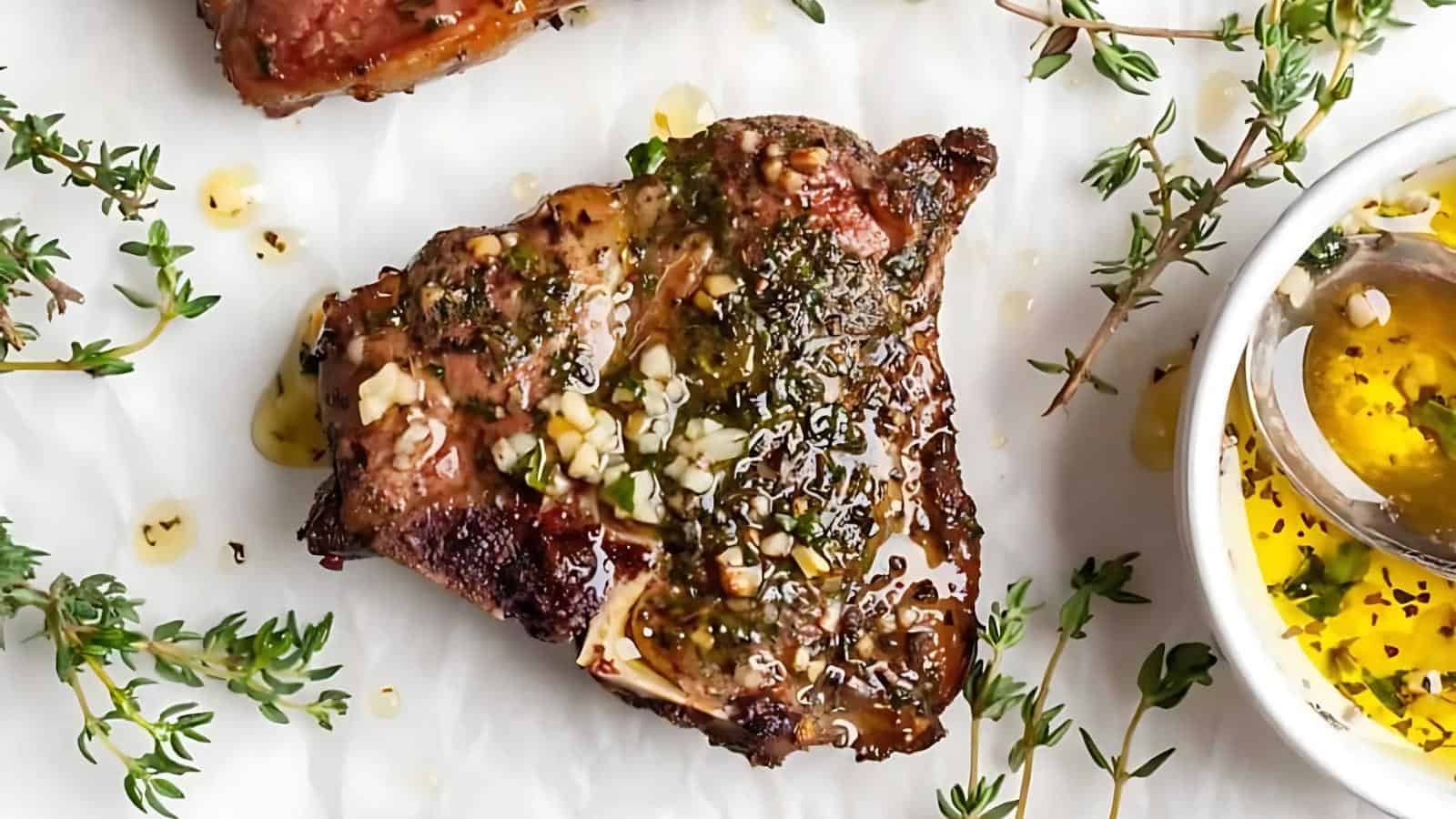 A cooked lamb chop topped with herbs and garlic is on parchment paper, surrounded by fresh thyme sprigs. A bowl of herb-infused oil is beside it.