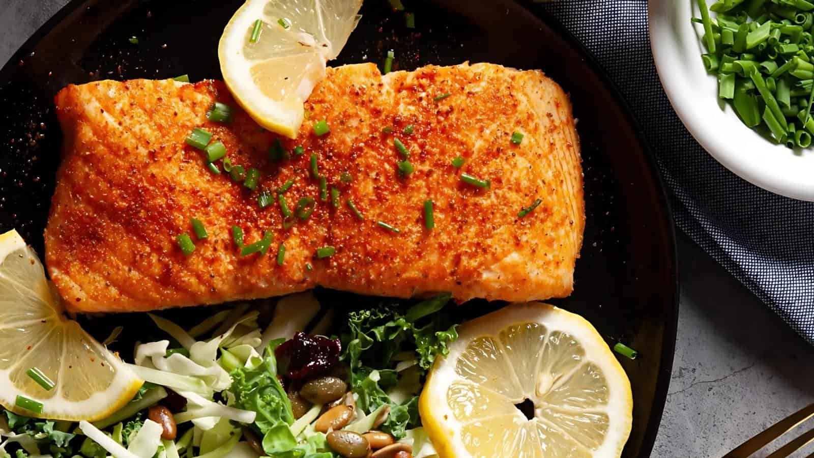 Grilled salmon fillet with paprika and chives, garnished with lemon slices, served on a plate with a side of leafy greens, cabbage, seeds, and cranberries.