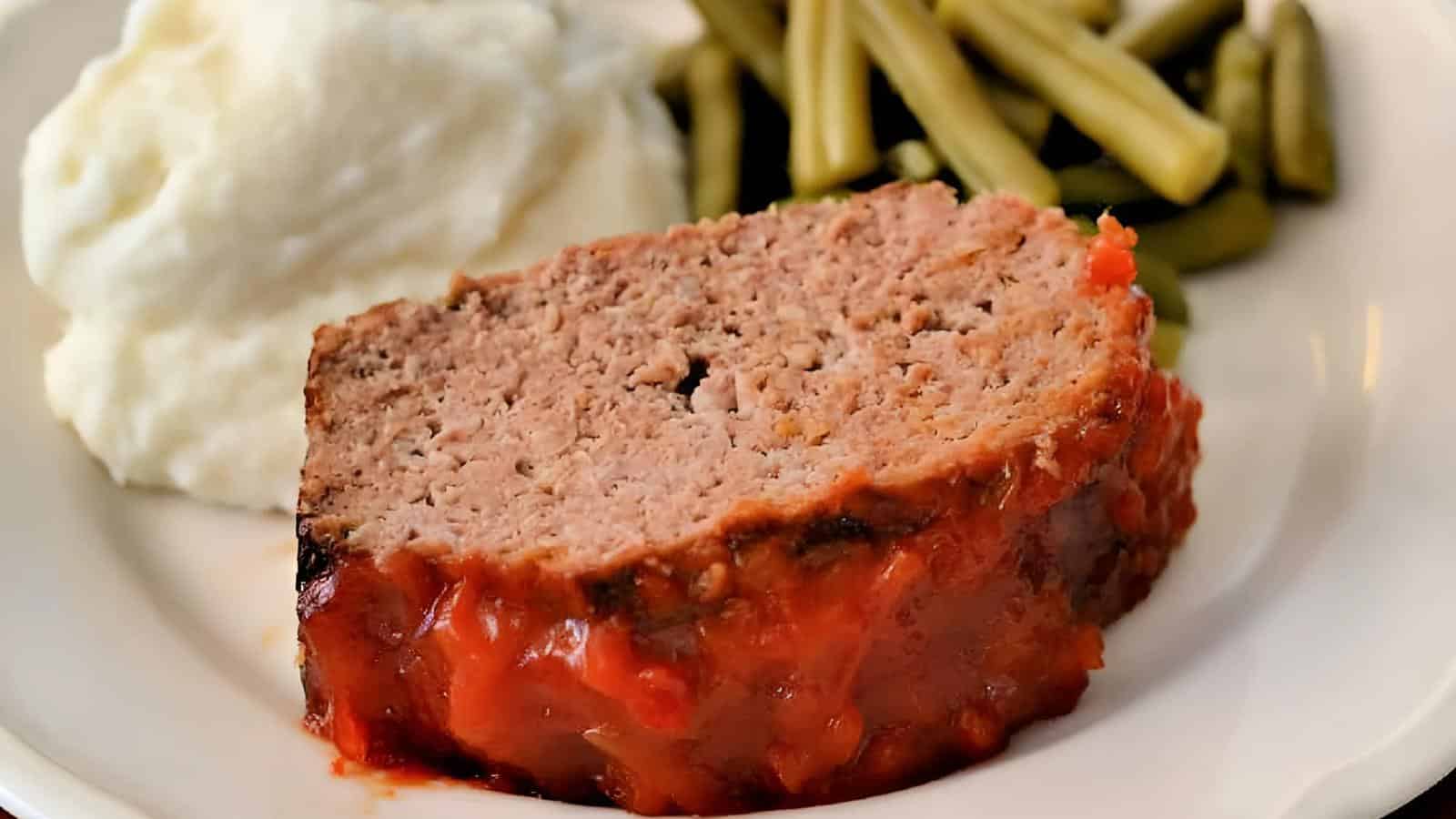 A slice of meatloaf with tomato glaze served on a plate alongside a portion of mashed potatoes and green beans.