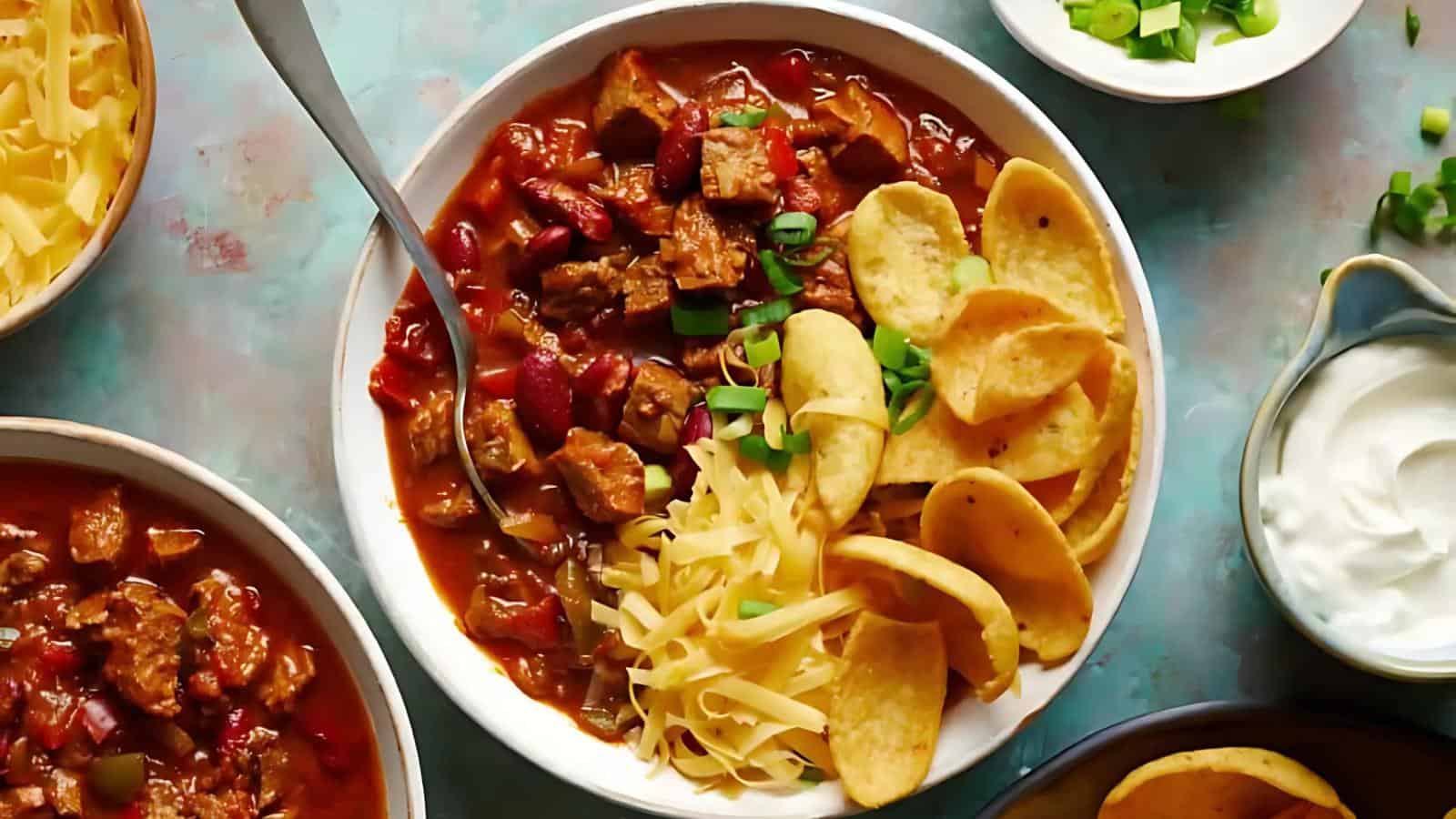 A bowl of chili with beef, kidney beans, cheese, and corn chips, garnished with green onions. Two smaller bowls contain shredded cheese and sour cream on the side.