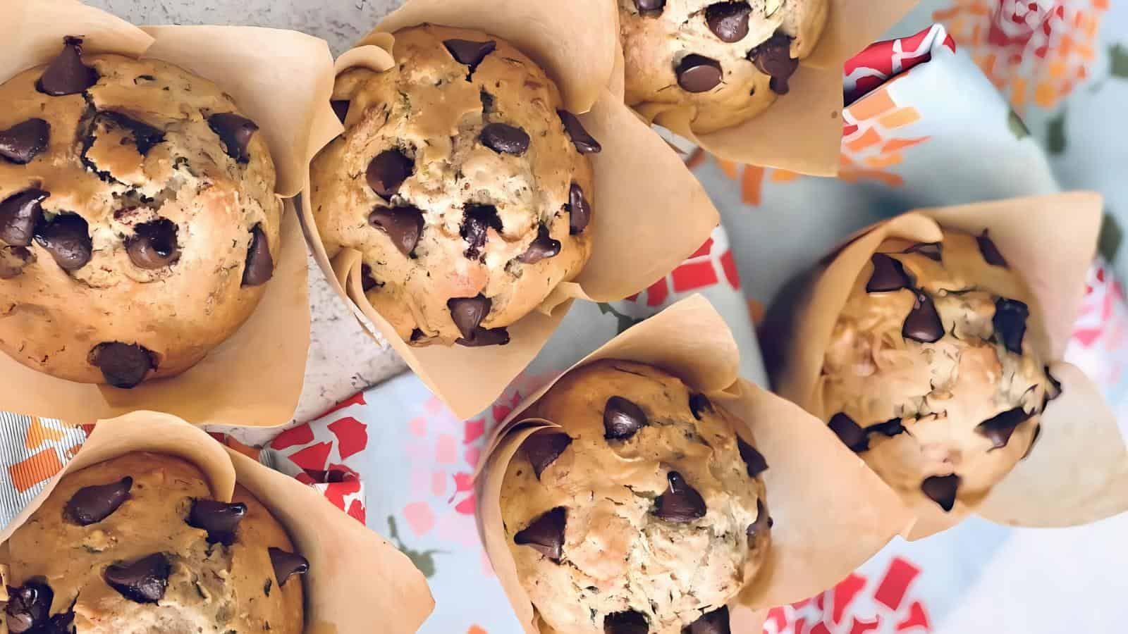 Six chocolate chip muffins in brown wrappers are arranged on a table with colorful paper underneath.