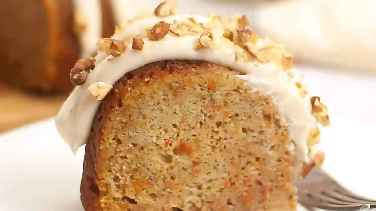 A slice of carrot cake with cream cheese frosting and walnuts on top, placed on a plate with a fork nearby.