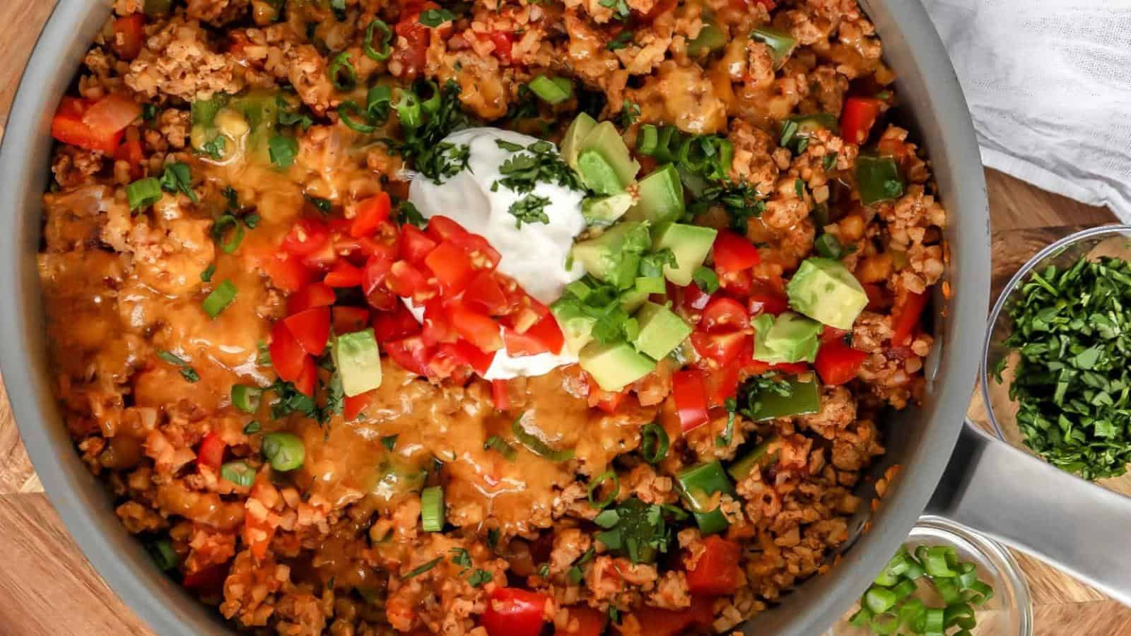 A skillet filled with a mixture of cooked ground meat, chopped tomatoes, avocado, and melted cheese, topped with a dollop of sour cream and garnished with green onions and herbs.